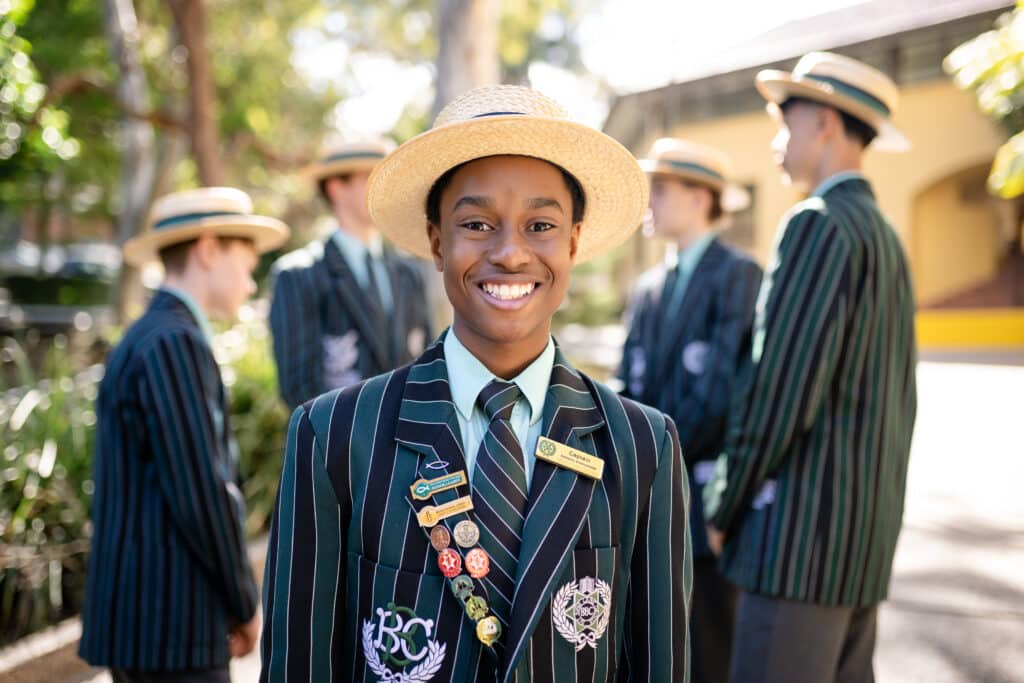 Nathan Maina in full school uniform with boater hat and blazer smiling at the camera. His blazer is adorned with multiple leadership and community badges.