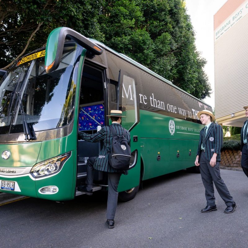 Three male students in school uniform with boater hat and blazer are lining up to step onto a green bus. The words 'More than one way to thrive' and the Brisbane Boys' College logo are printed on the side of the bus.