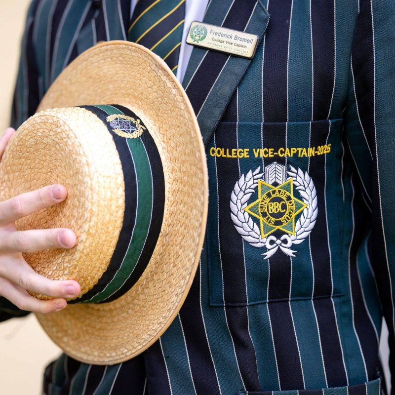 A photograph of a Brisbane Boys' College blazer and the BBC boater hat. The BBC crest is visible with the motto 'Sit sine labe decus'. Above the crest are the words 'College Vice-Captain 2025' in yellow stitching.