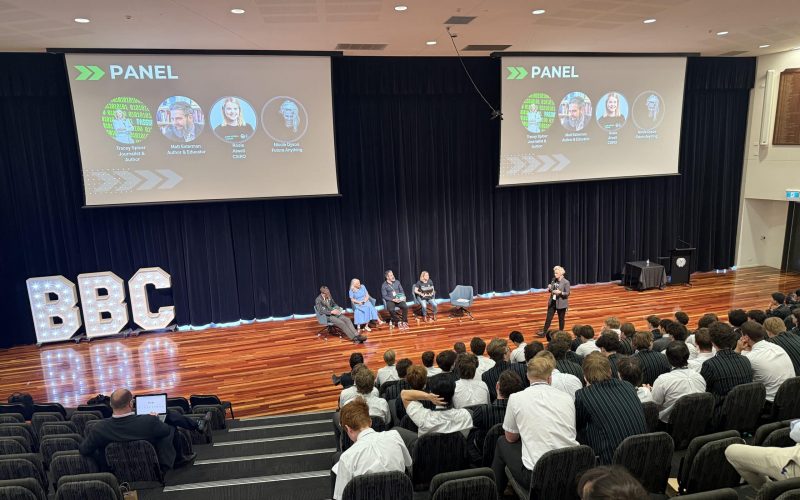 Students seated inside an auditorium in front of a panel of five adults.